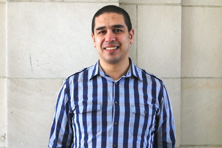 Visiting Assistant Professor of English Karim Ibrahim, with short dark hair in blue-and-brown plaid business shirt.