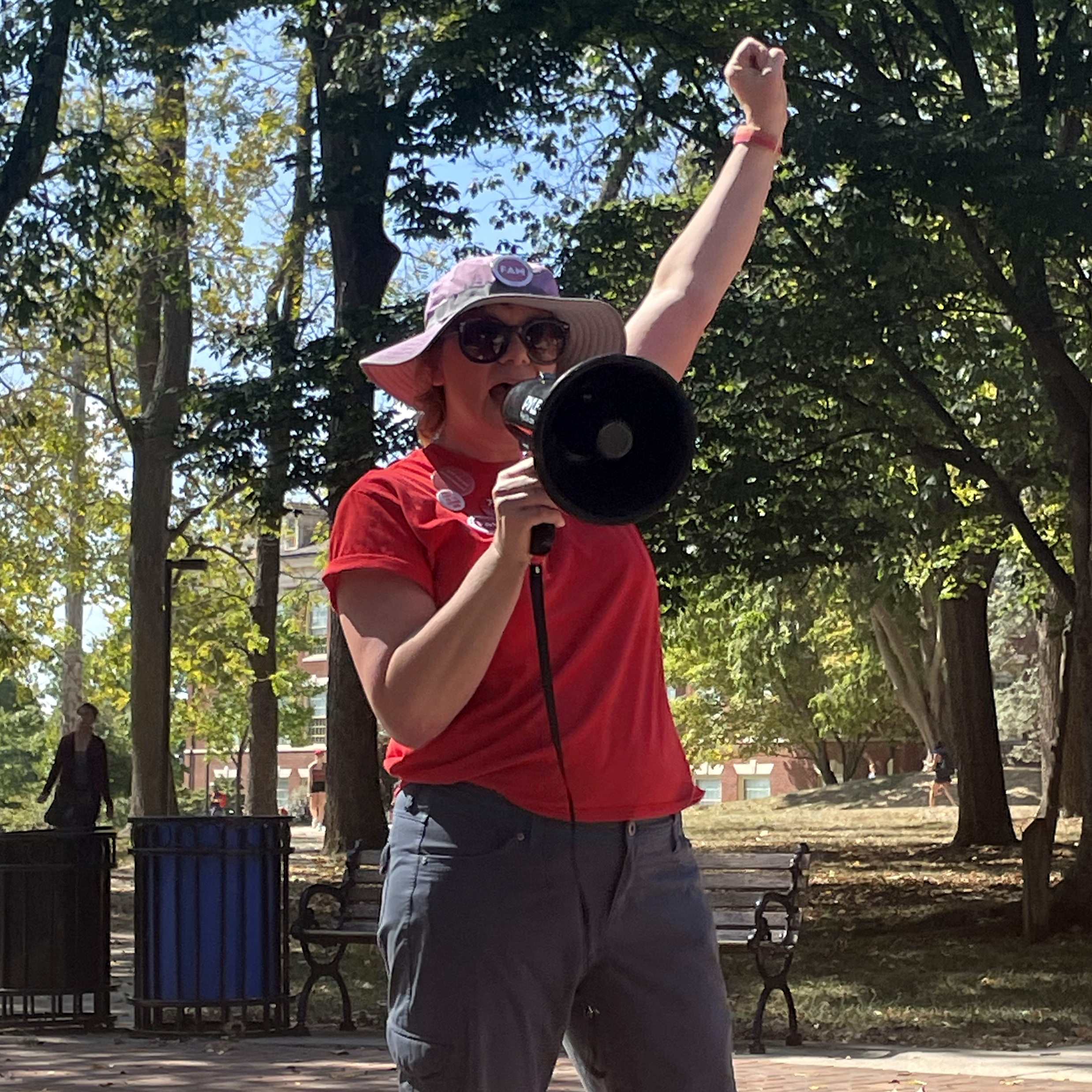 Theresa Kulbaga, co-chair of FAM's Negotiating Team, wearing a red FAM t-shirt and a hat, raises her fist while speaking into a megaphone at FAM's Rally for Raises at the tree-lined Seal on Miami's Oxford Campus